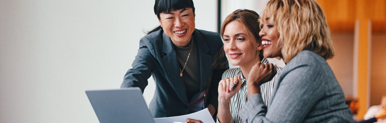 Three female colleagues are sitting in front of a computer talking and smiling.