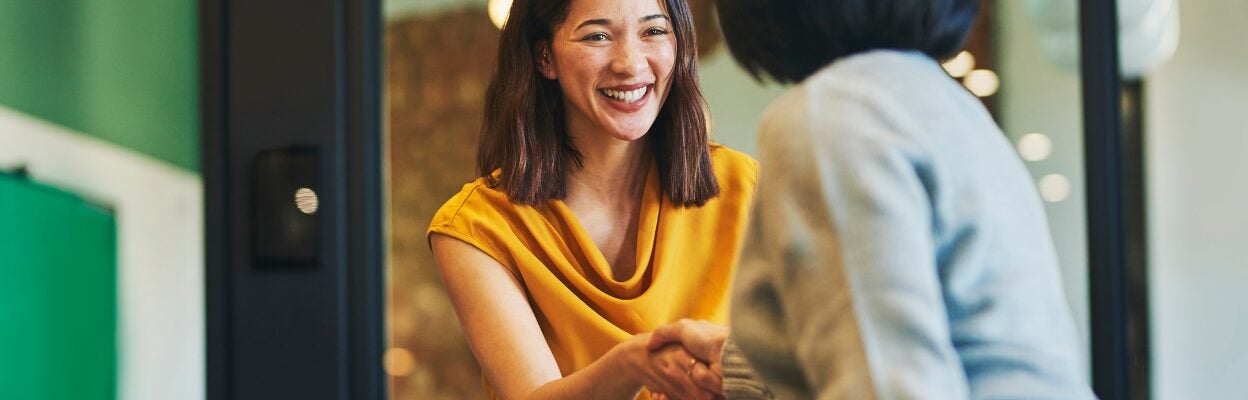 Two female colleagues meet for the first time. They are shaking hands and smiling while standing at a conference table in a modern office.
